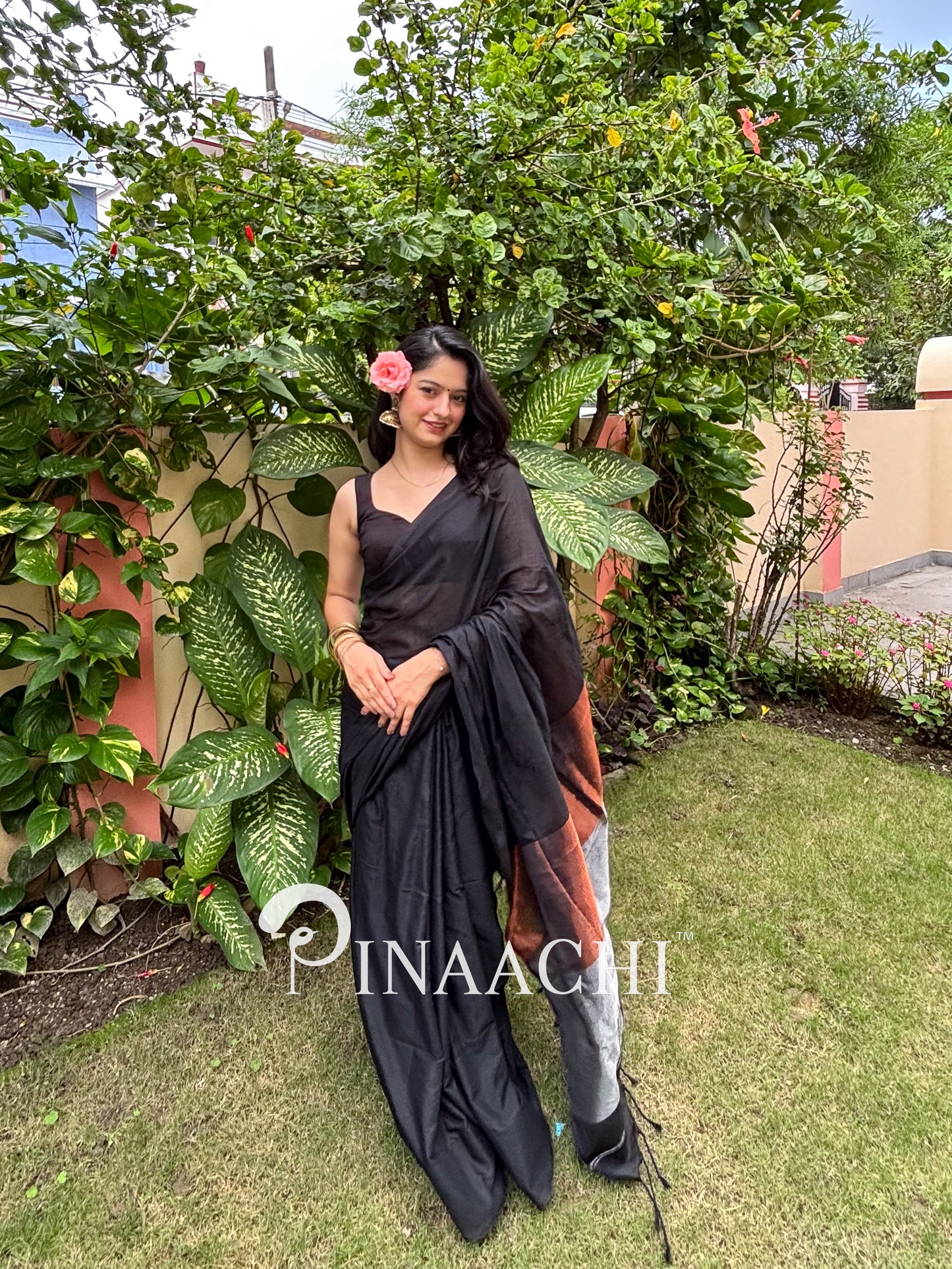 Model wearing a black cotton saree with zari border, set against a lush green background