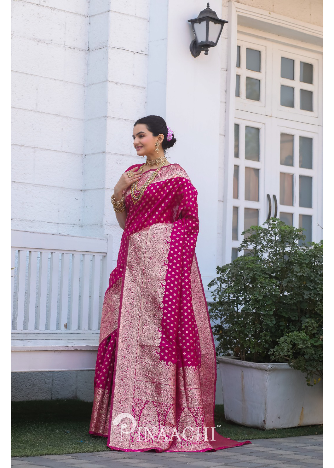 Woman wearing Pinaachi Pink Banarasi Silk Saree with intricate gold patterns standing outdoors near white building