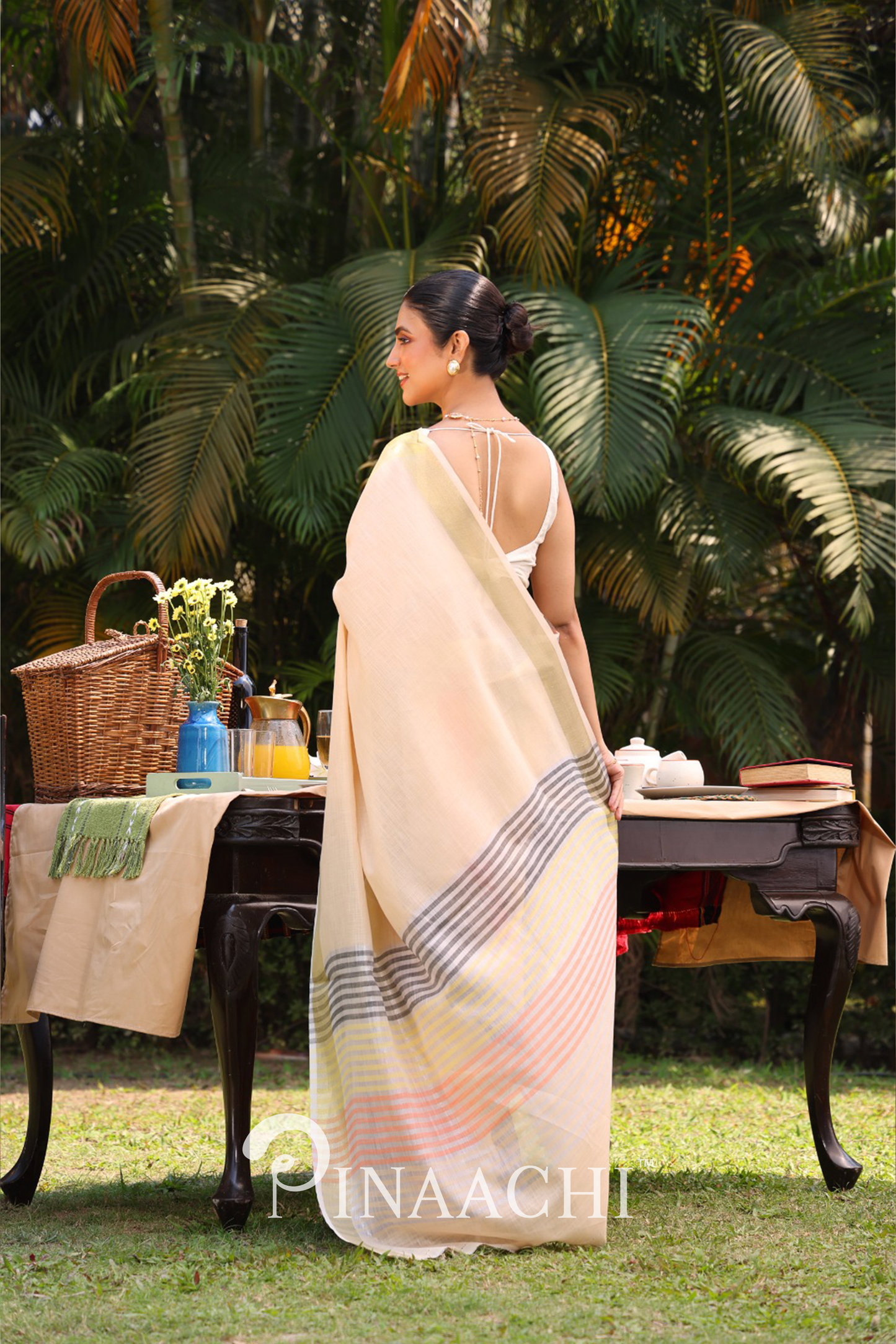 Woman wearing Pinaachi striped linen silk saree standing outdoors near wooden table with picnic items