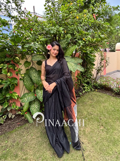 Model wearing a black cotton saree with zari border, set against a lush green background