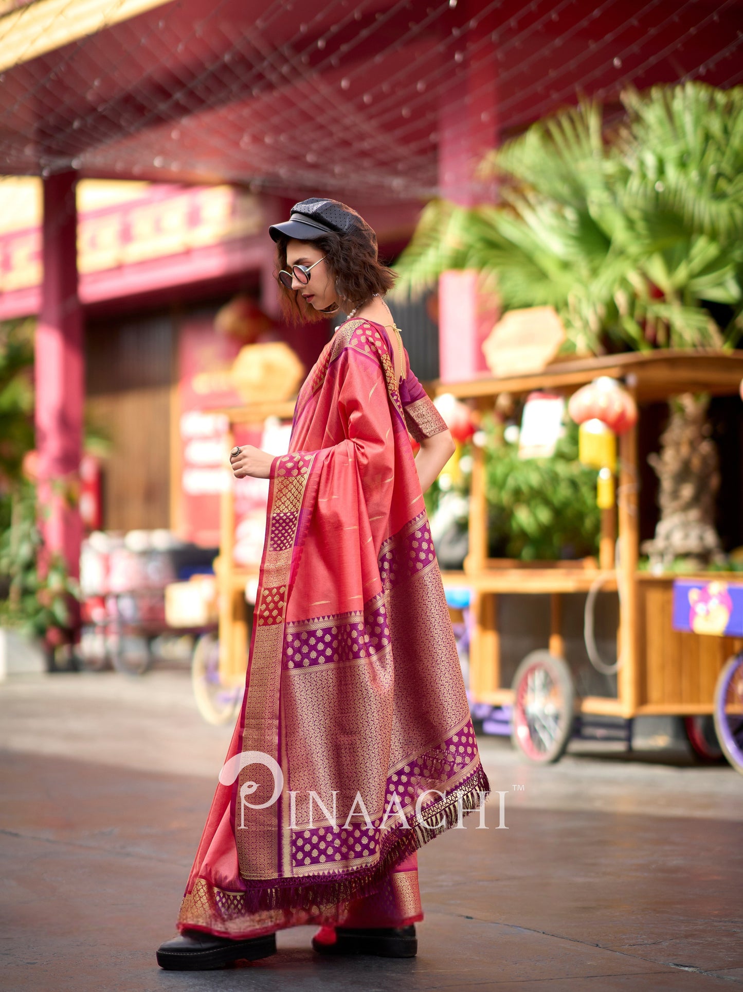 Stylish model wearing a coral Banarasi silk saree with intricate gold patterns at a vibrant outdoor setting