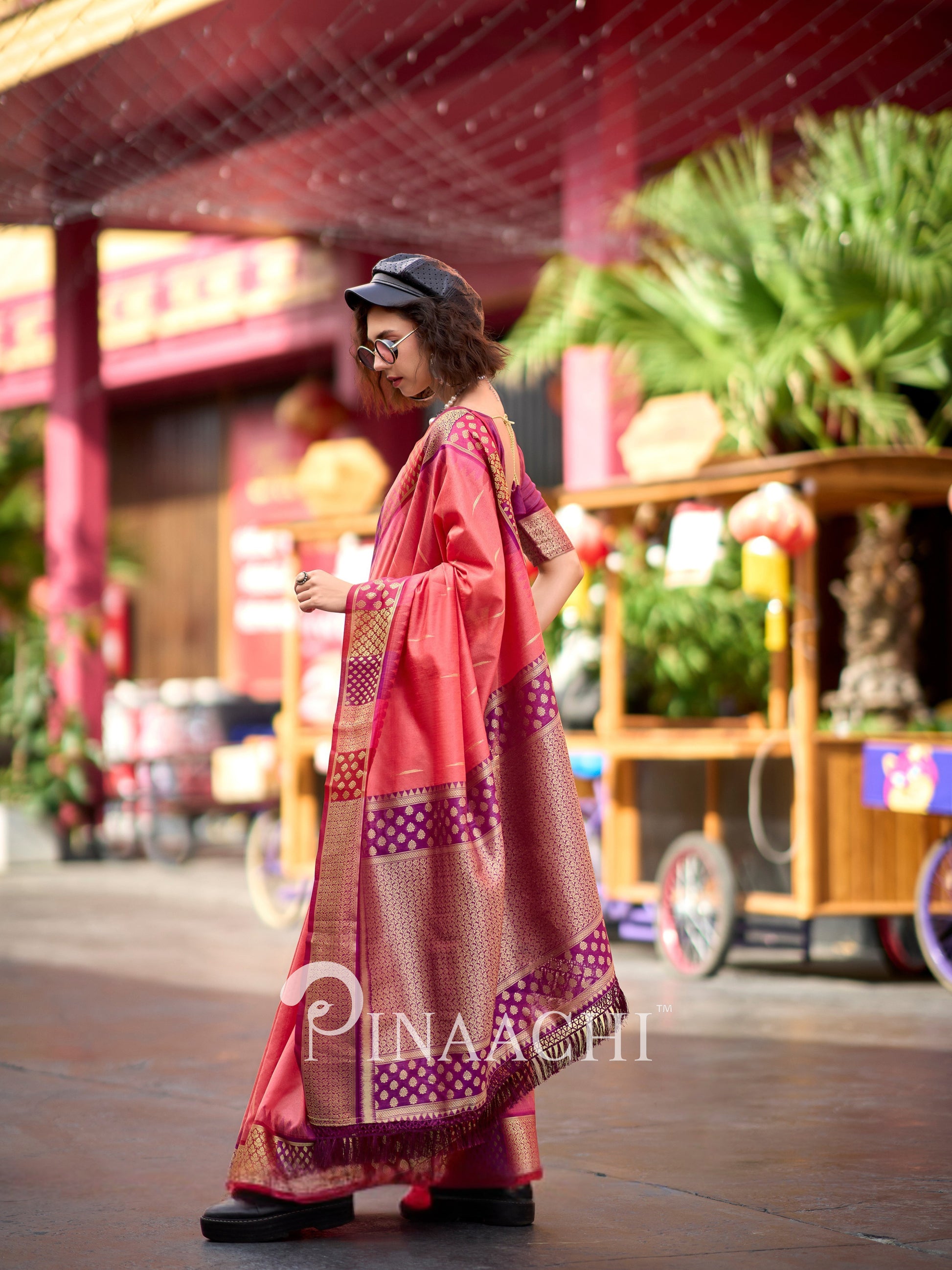 Stylish model wearing a coral Banarasi silk saree with intricate gold patterns at a vibrant outdoor setting