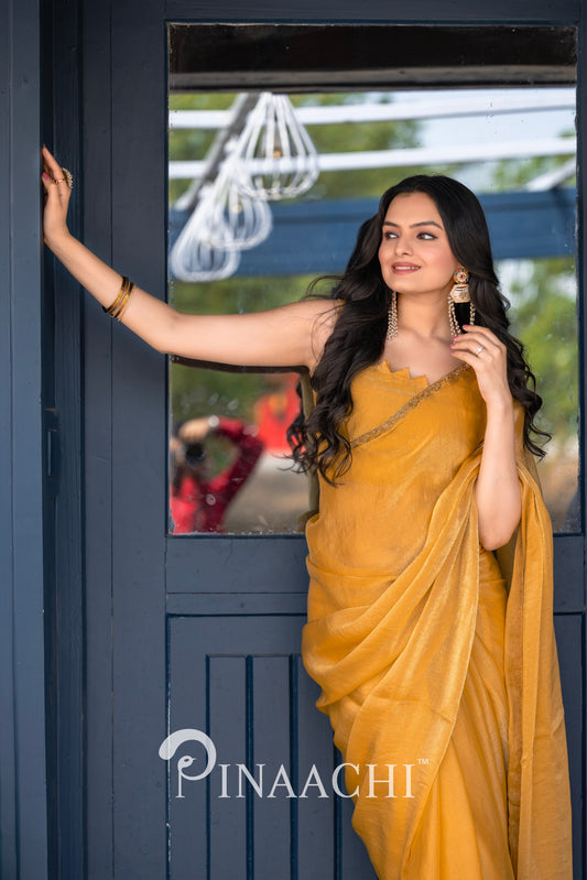 Woman wearing pinaachi solid yellow chiffon saree standing by a door with long curly hair and gold earrings