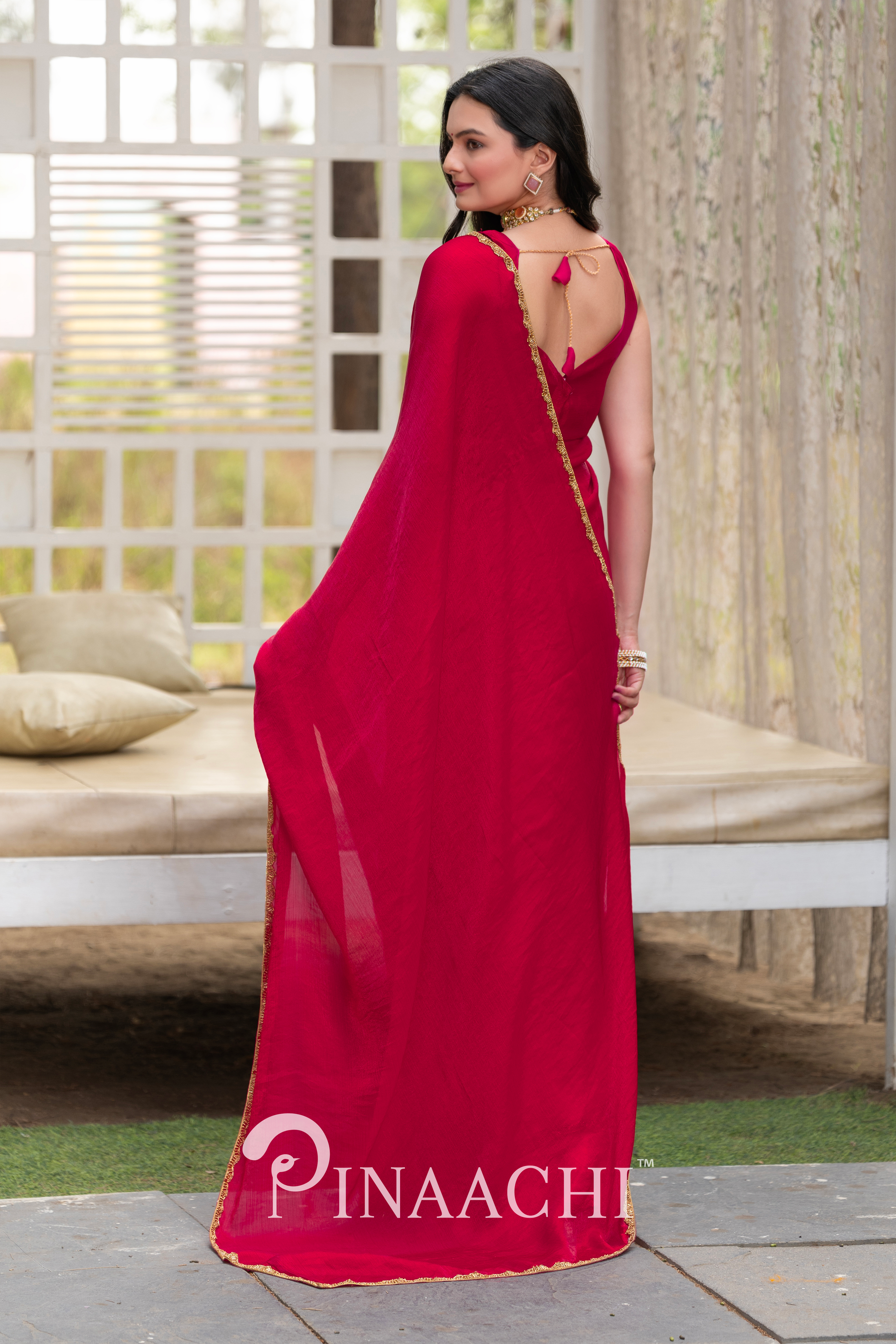 Woman wearing Pinaachi solid cherry red chiffon saree with gold border posing indoors
